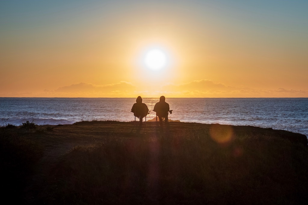 2 persons seated in chairs watching the sunset over a sea