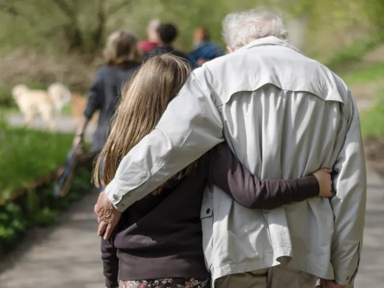 Photo of a young girl on the left and old man on the right, walking along a paved trail in a park, each with an arm around the other.