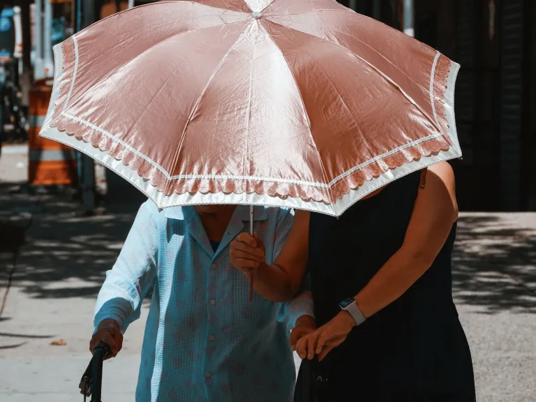 Photo of two women walking under a parasol; an elderly woman with a walker being helped by a middle-aged woman.