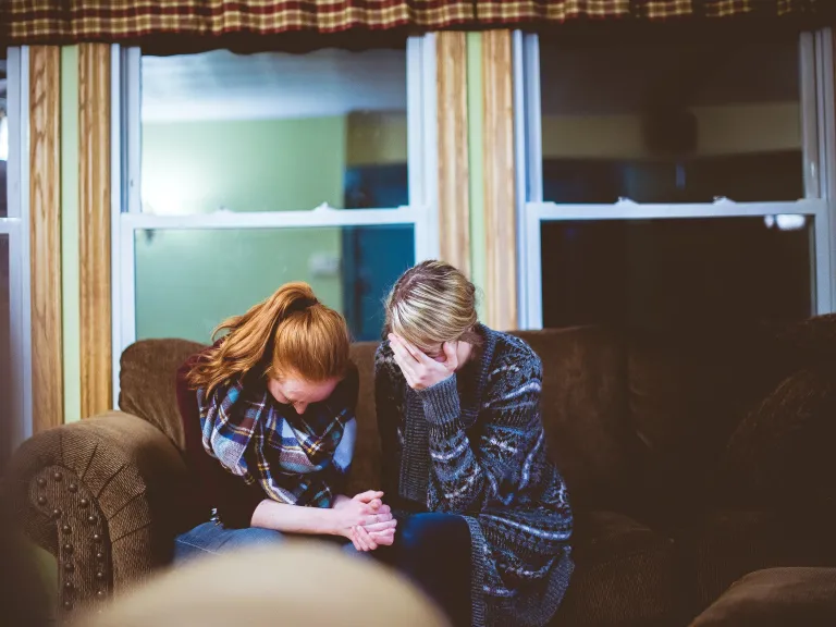 Photo of two women sitting on the couch grieving together. One woman has her hands clasped and one woman is leaning her face into her forhead.