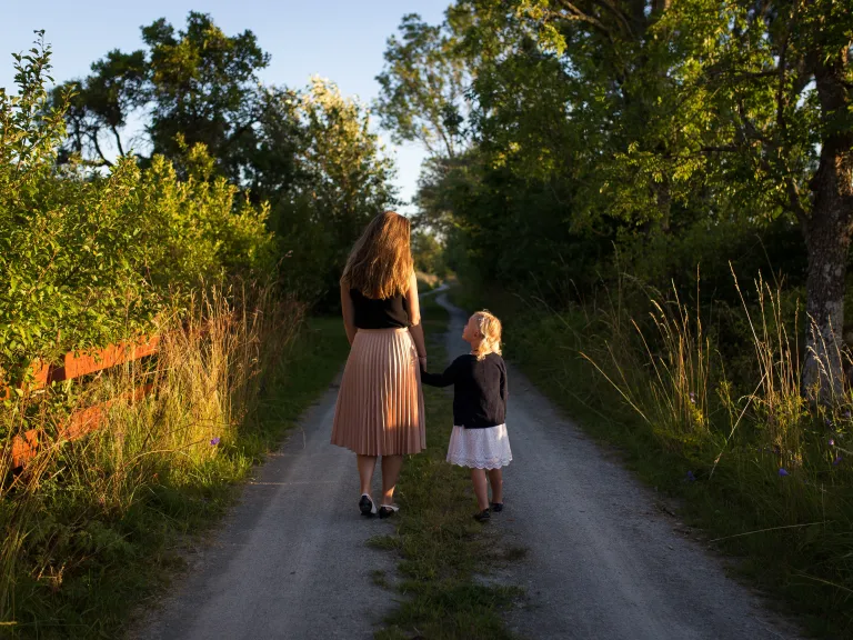 Photo of a mother and young daughter walking down a dirt road in the countryside; the daughter is holding her mother's hand and looking up at her.