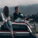 Two young adult women sitting on top of a car.